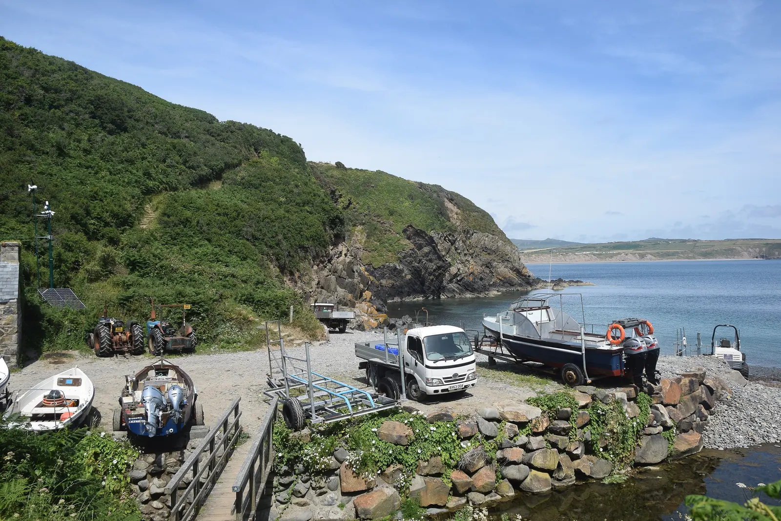 Lobster boats, tractors and gear drawn up on the shingle slipway at Porth Meudwy, the cove opening to the sea beyond