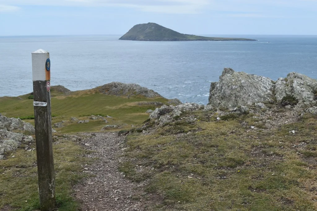 A weathered waymark post on the clifftop coast path above Aberdaron, with Ynys Enlli lying across the Sound