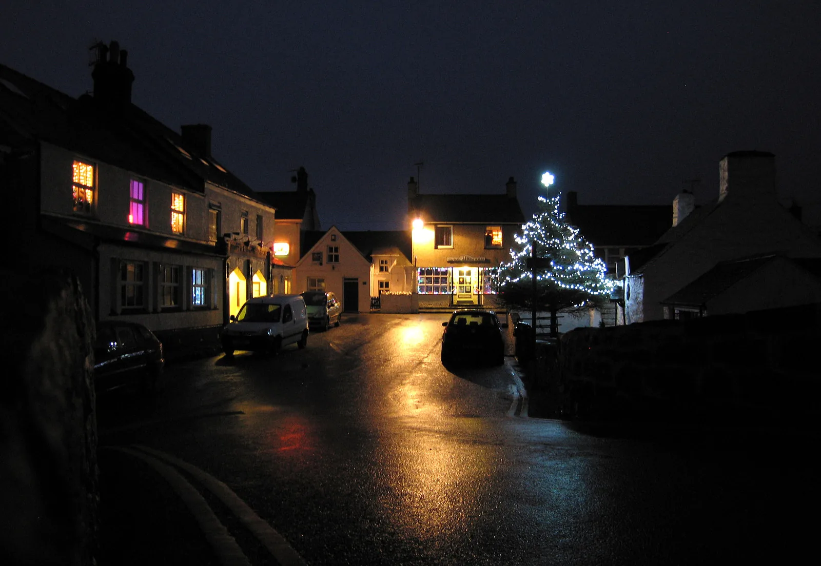 Aberdaron’s street at night in winter, lit windows and a lit tree reflected on the wet road
