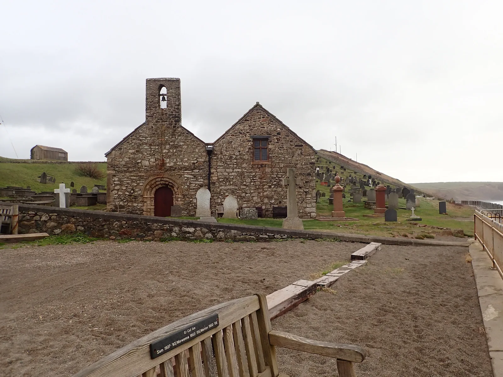 St Hywyn&rsquo;s churchyard at Aberdaron under a grey sky, headstones running down towards the sea wall and the bay beyond