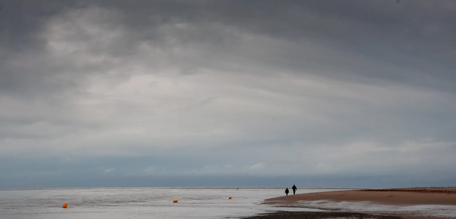 Wide empty sand and the Dyfi channel under a grey sky, two walkers in the distance