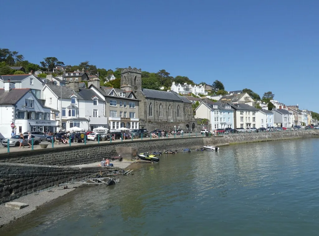 Aberdyfi&rsquo;s seafront seen from the water in summer, St Peter&rsquo;s Church tower rising above the terraces and people along the sea wall