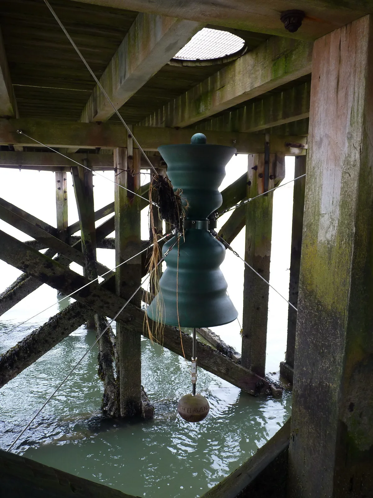A bronze bell hanging in the timber framework beneath Aberdyfi jetty, just above the water