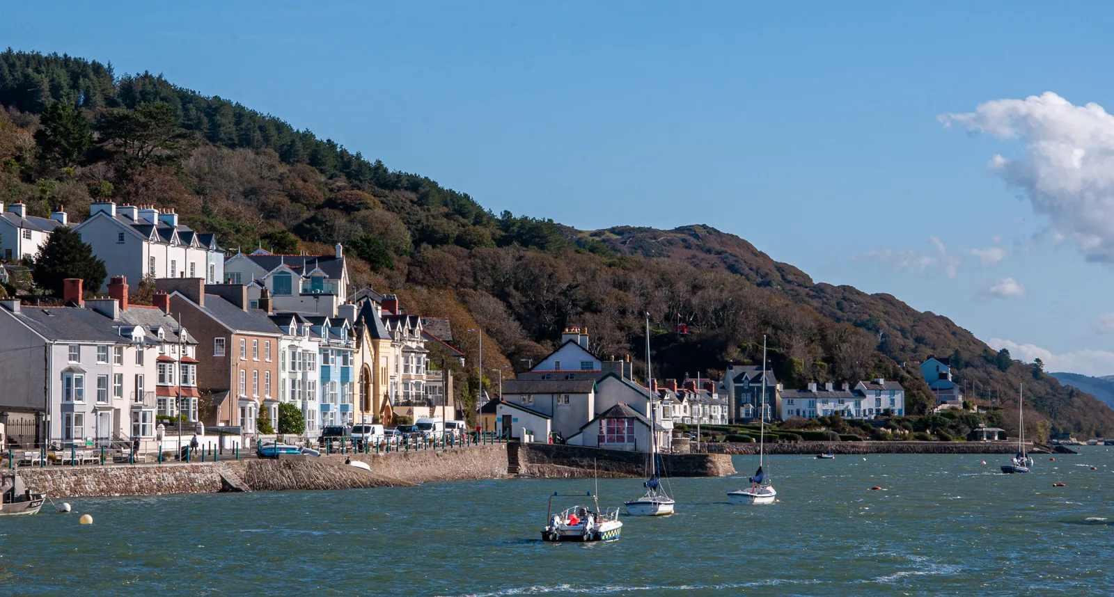 The painted seafront terraces of Aberdyfi below a wooded hillside, with moored boats on the Dyfi estuary in front of them