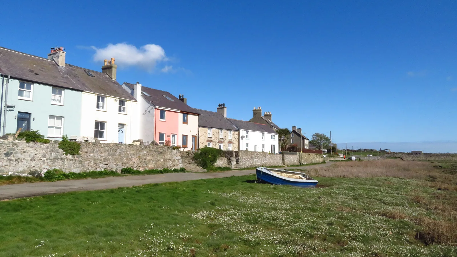 A row of pastel-painted cottages at Aberffraw standing above the grass and the tidal Afon Ffraw, a small boat drawn up on the bank