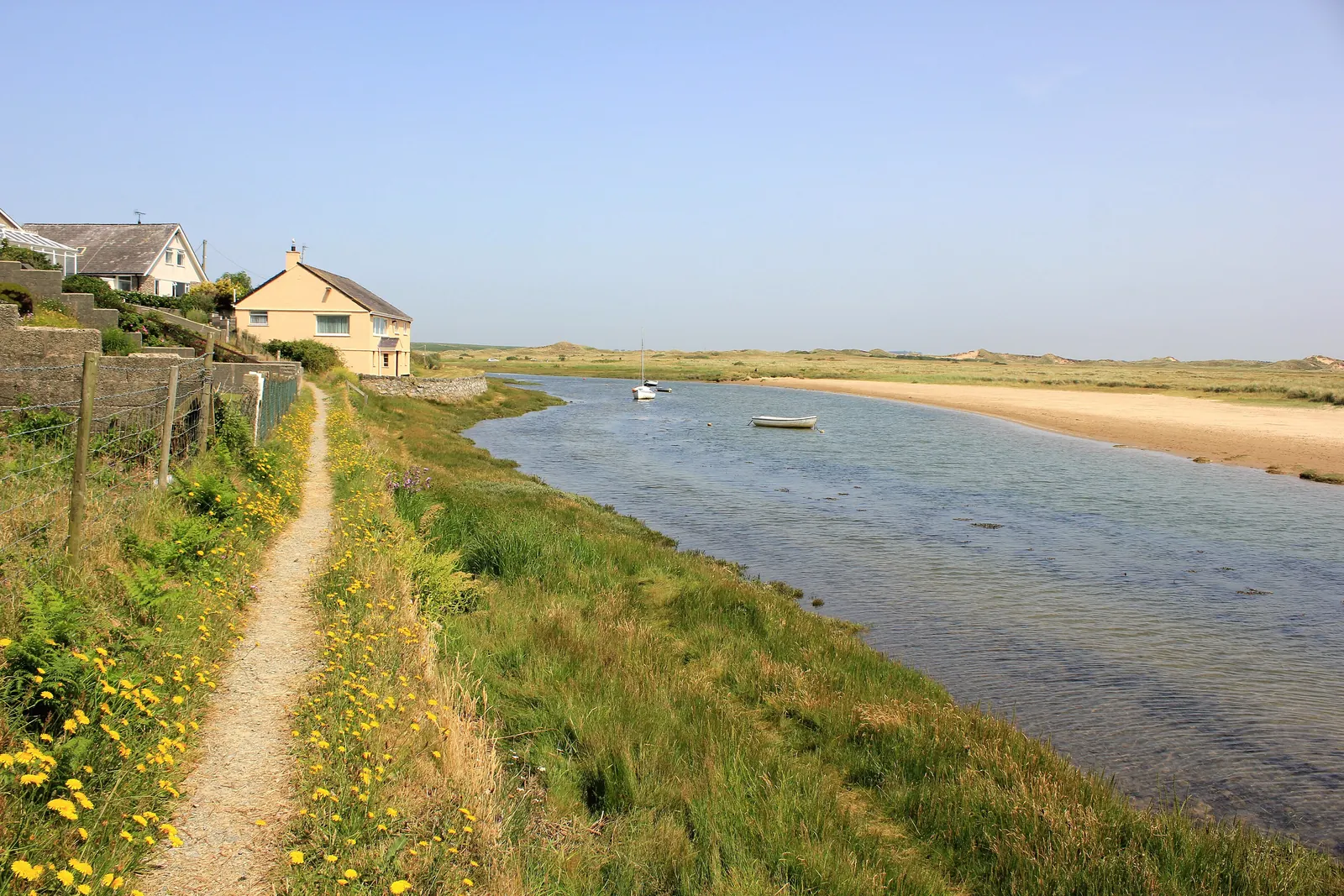 The Afon Ffraw estuary at Aberffraw opening out towards the sea, the coast path running down past the village houses with a boat moored on the water
