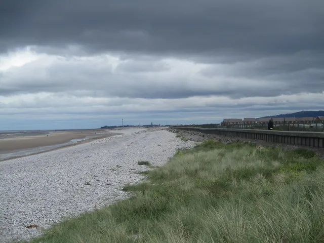 The shingle ridge at Traeth Pensarn, marram and sea plants along its crest, with the promenade and the caravan park behind and the bay opening west