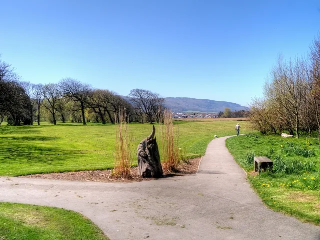 A surfaced path curving through Pentre Mawr Park, Abergele, past a dark metal sculpture with the hills behind