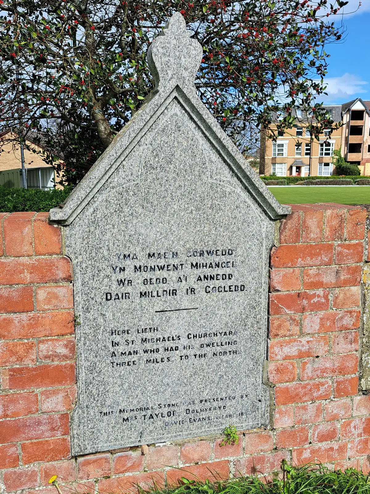 A pale gabled memorial stone set against a brick wall in St Michael&rsquo;s churchyard, Abergele, its inscription recording a man who lived three miles to the north