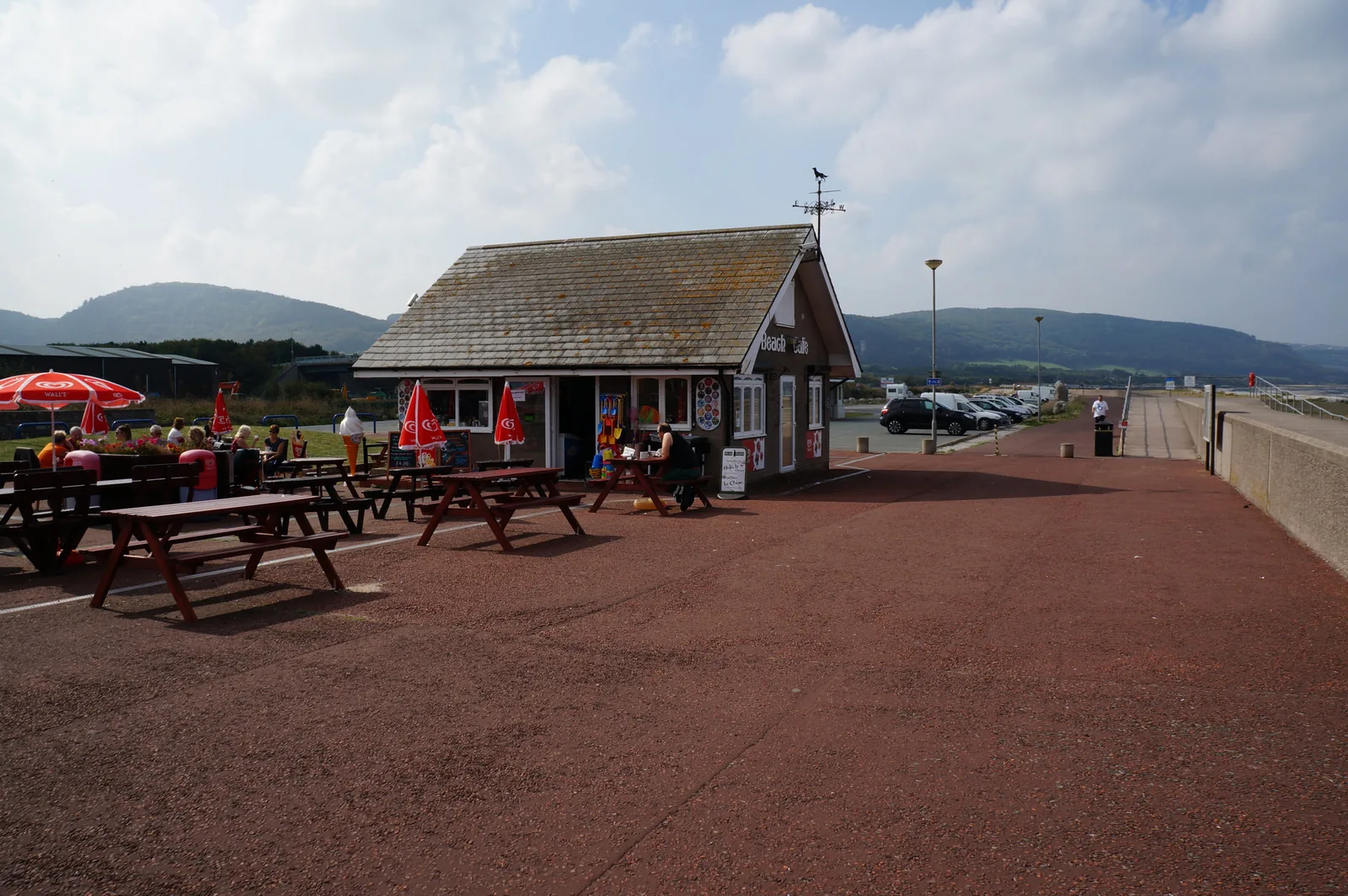 Picnic benches and parasols outside the beach caf&eacute; on Pensarn promenade, the red tarmac path running away towards the hills