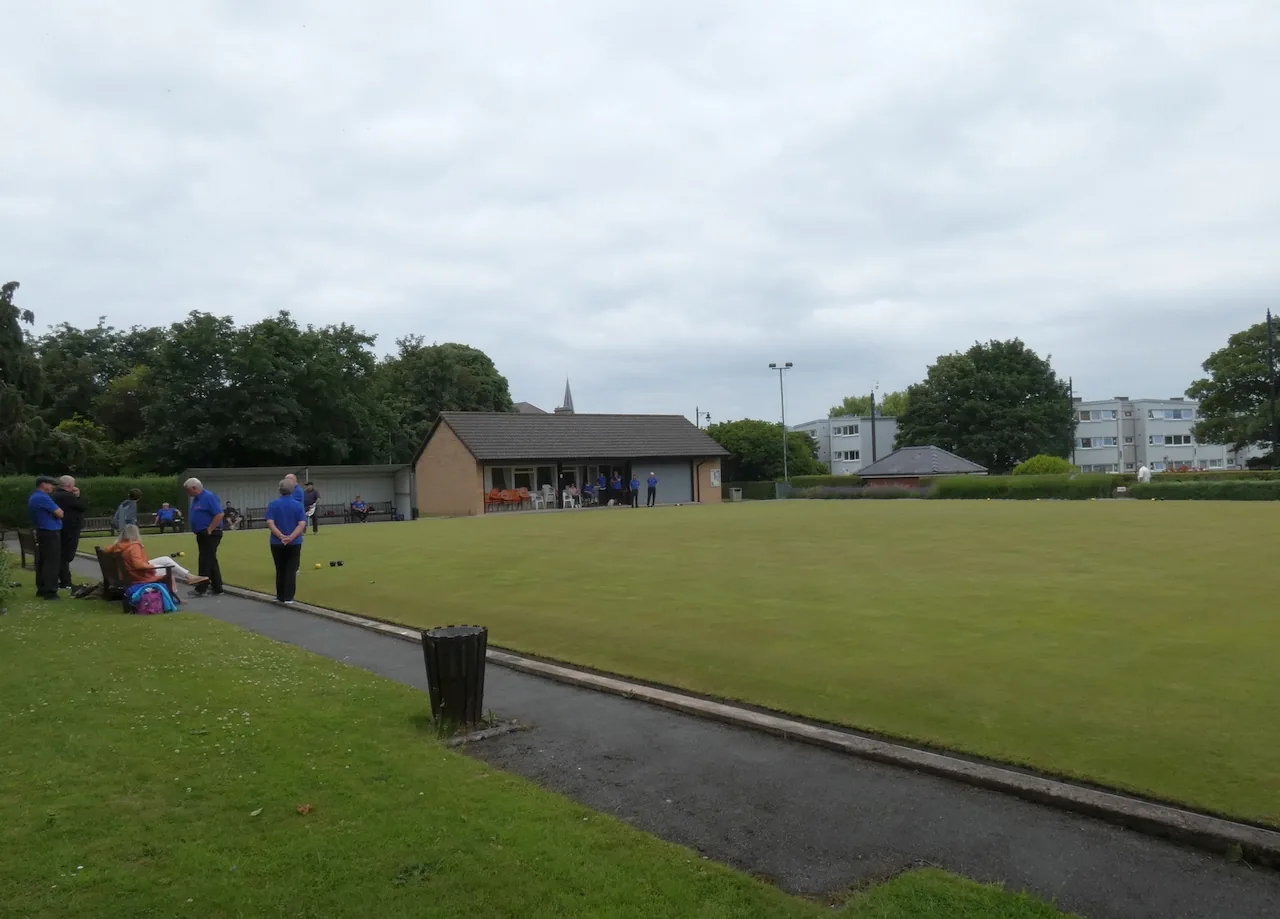 Players in white on the flat green of Abergele bowling green, the pavilion and rooftops behind