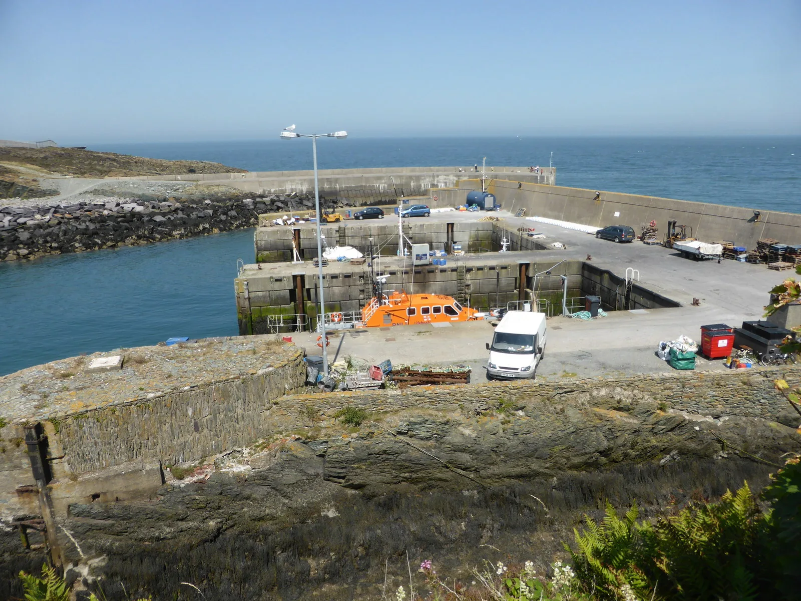 The dry docks and breakwater at Porth Amlwch seen from above, an orange lifeboat on the quay and open sea beyond