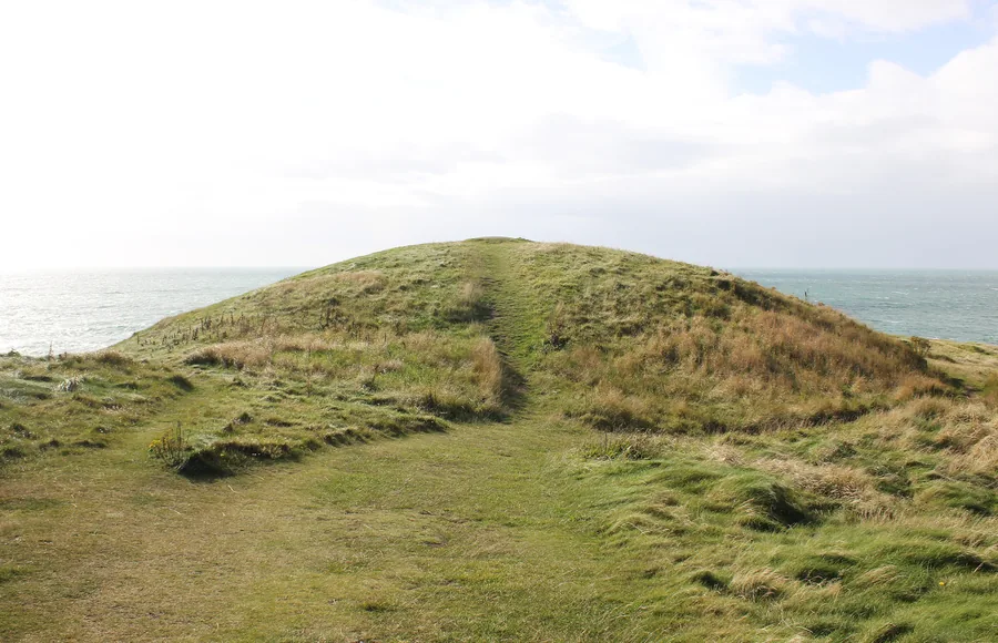 The turf-covered mound of Barclodiad y Gawres on its headland above Porth Trecastell, a worn path running over it and open sea on both sides.