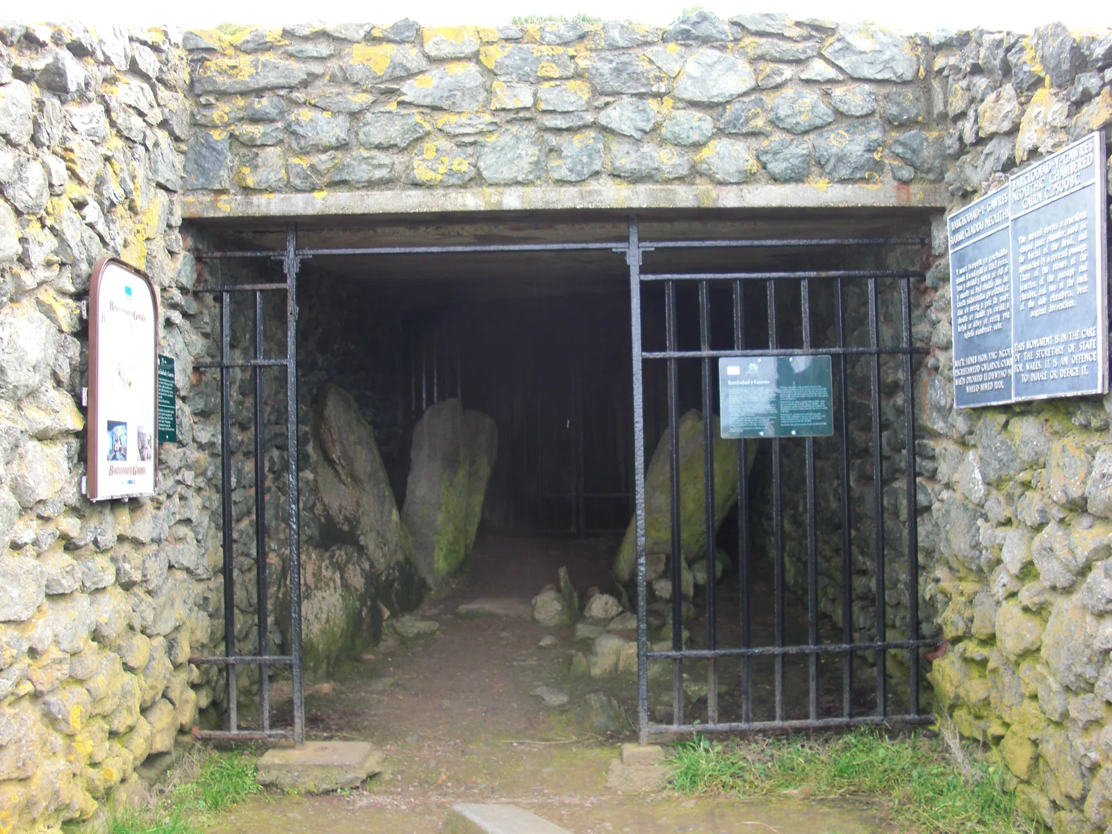 The barred entrance gate of Barclodiad y Gawres set in its stone forecourt wall, with two Cadw interpretation boards on either side.