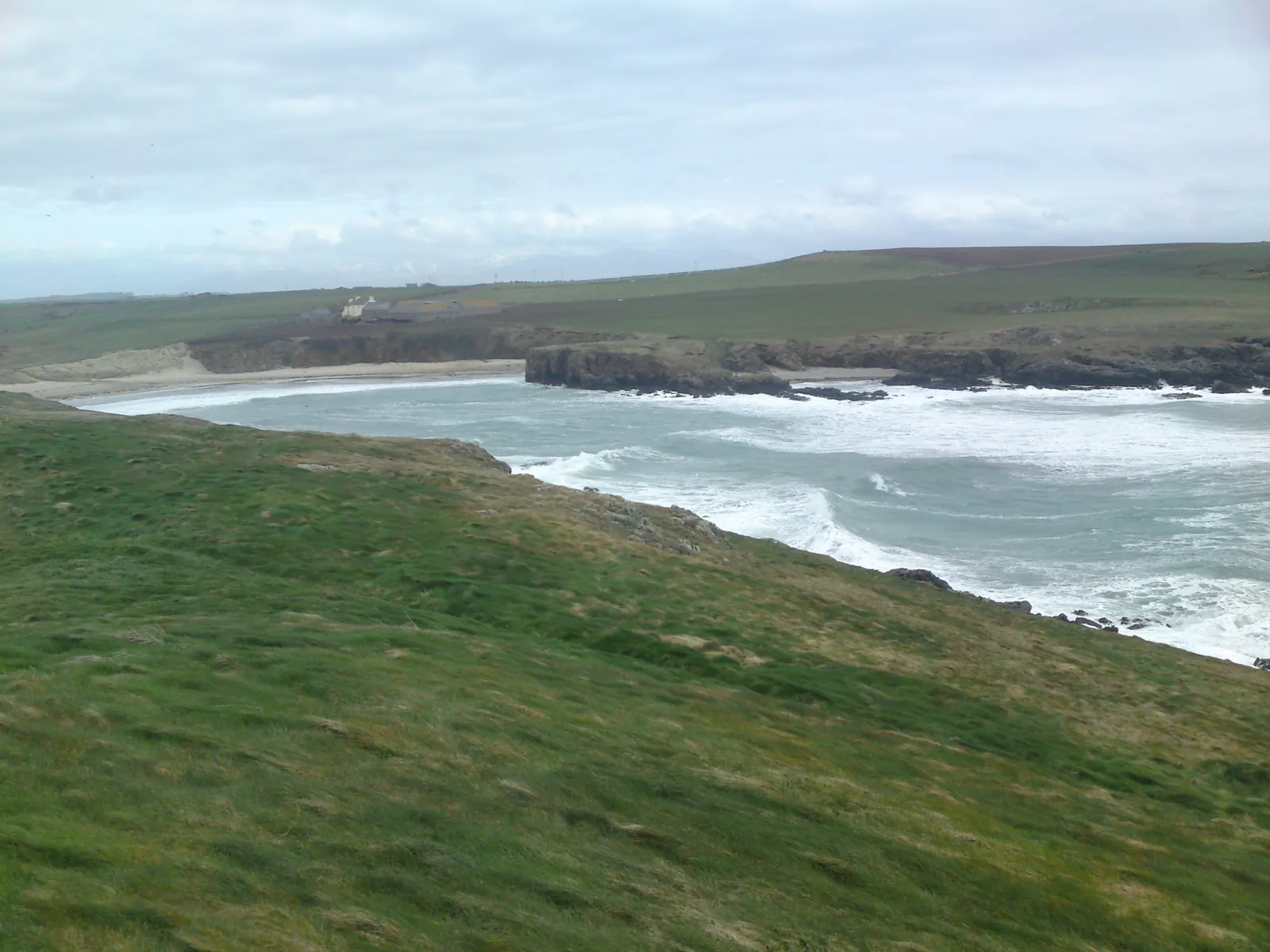 Porth Trecastell seen from the headland at Barclodiad y Gawres, surf running into the sandy cove between low cliffs.