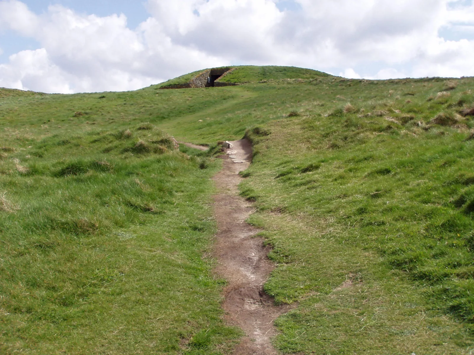 A worn earth path climbing over grass toward the mound of Barclodiad y Gawres, its stone-framed entrance visible in the flank at the top.