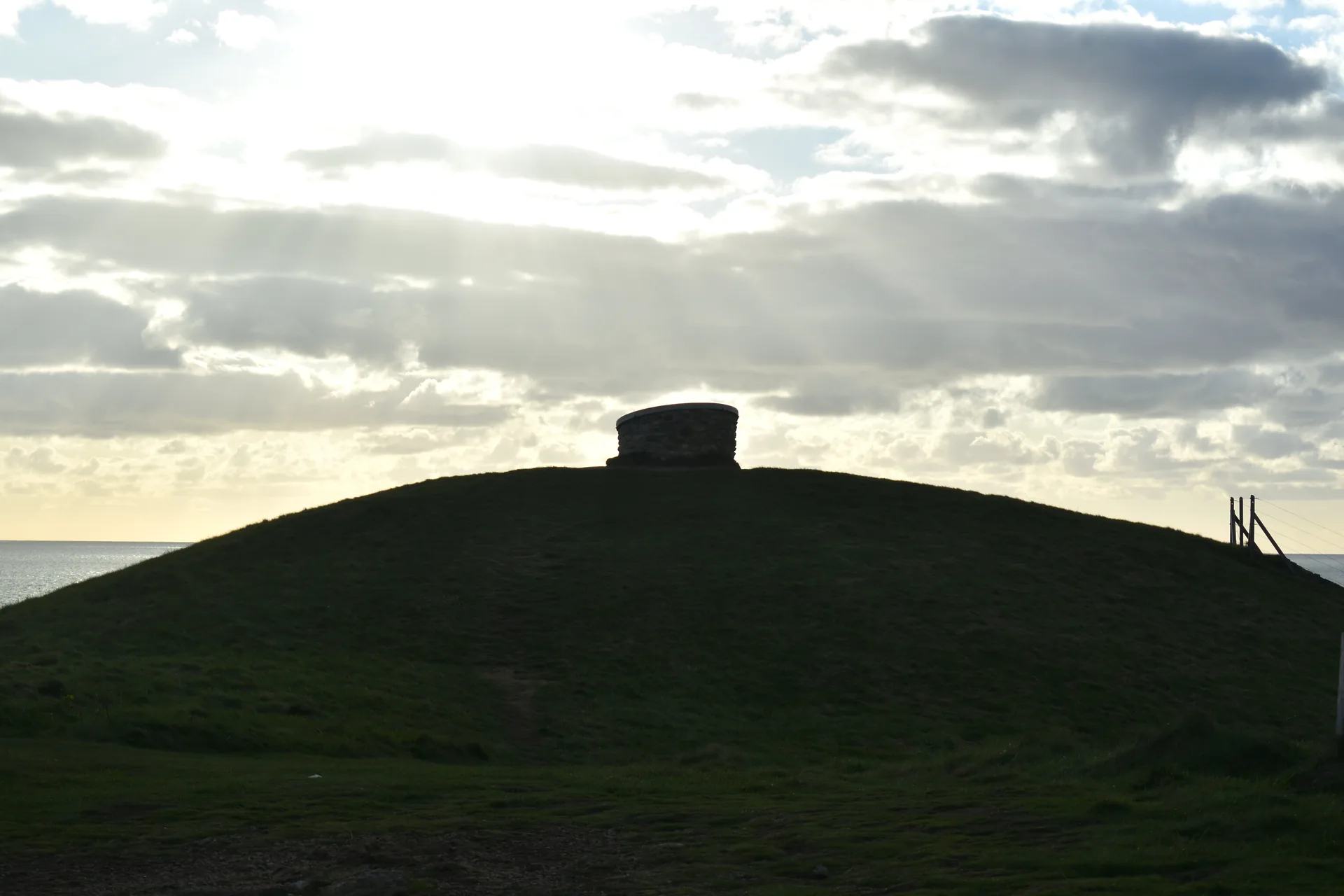 The turf-covered mound of Barclodiad y Gawres silhouetted on its headland against a bright broken sky, the sea beyond and the stone roof-light on its crown.