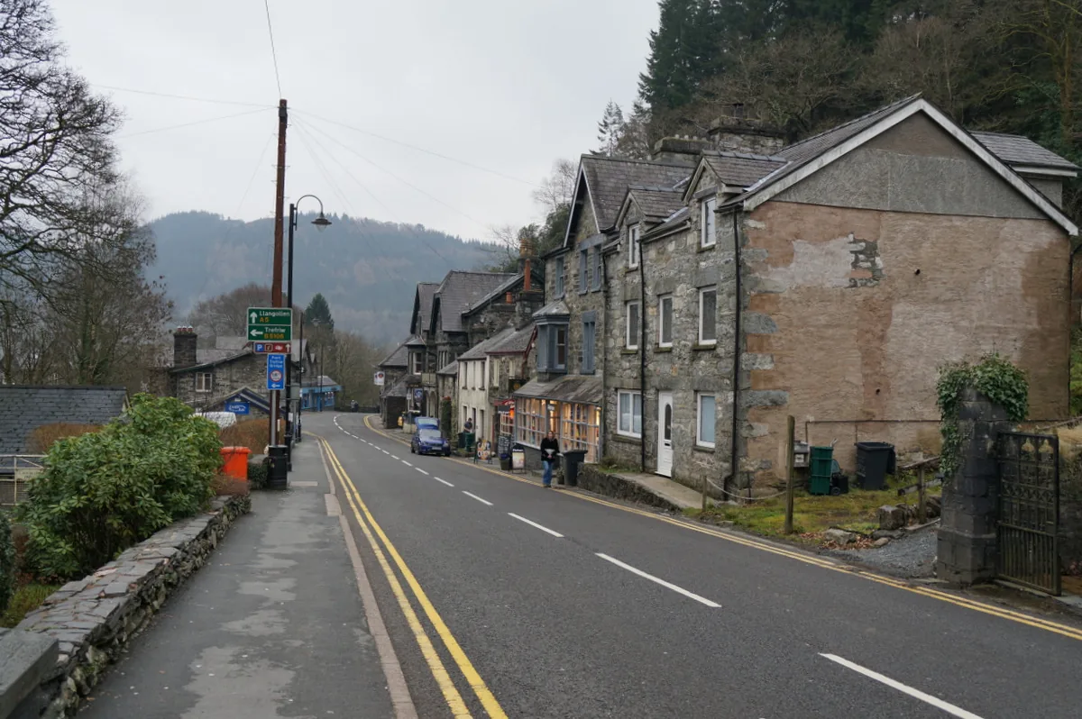 The main road through Betws-y-Coed, with stone buildings, a lit shopfront and wooded hillside above