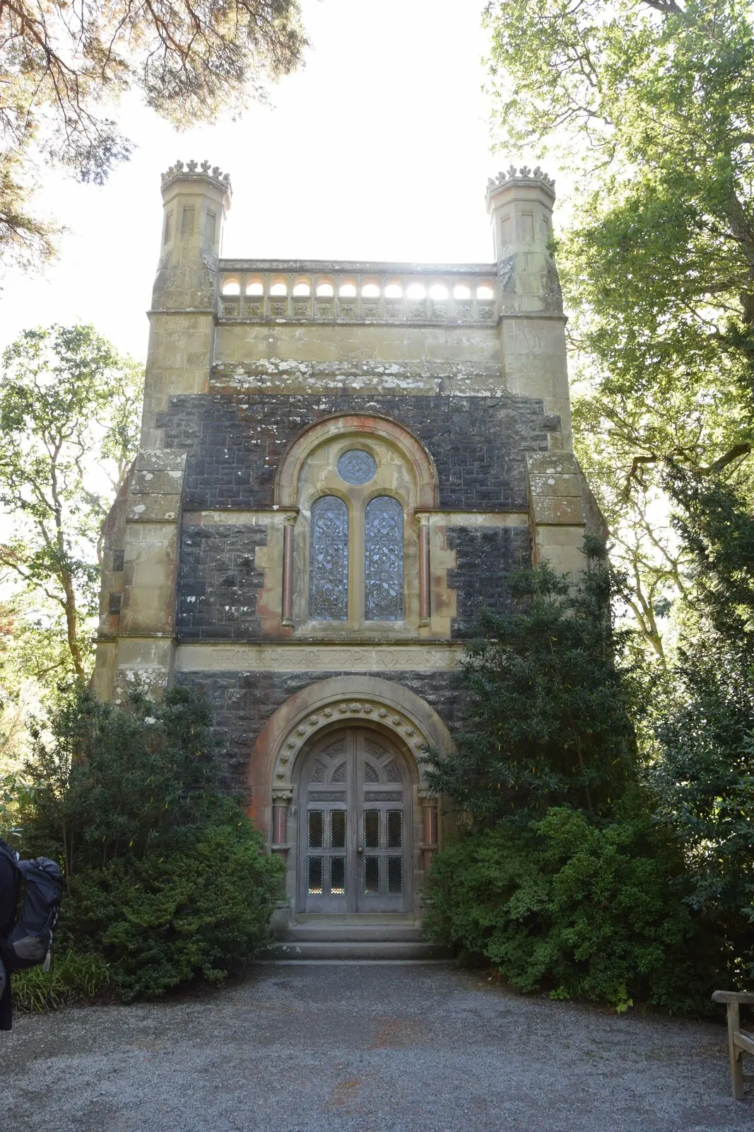 The Poem, a Gothic-arched stone mausoleum at Bodnant Garden, framed by trees at the end of a gravel path.