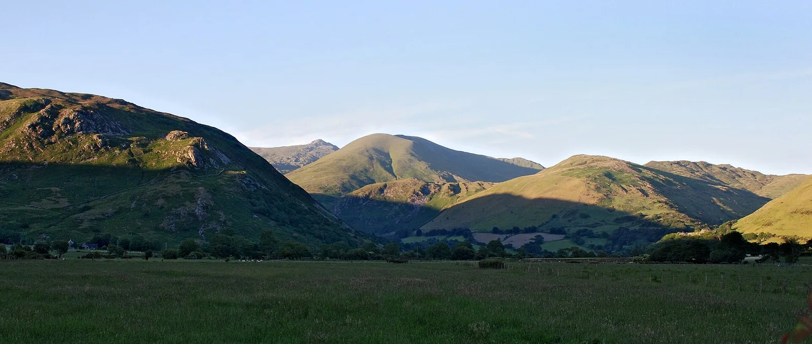 Low golden evening light raking across the layered ridgelines and cwms of the Cadair Idris massif in southern Eryri, Gwynedd, above a broad green valley.