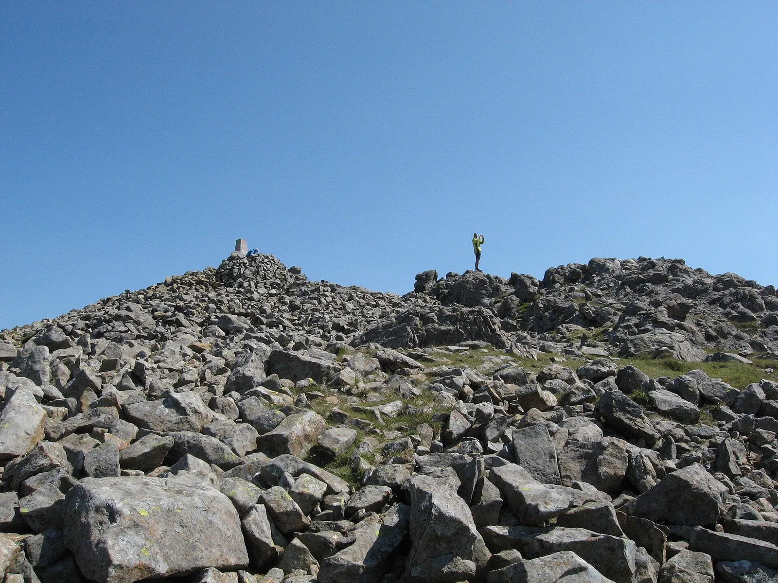 The rocky summit of Cadair Idris (Penygadair) on a clear day: the trig point on its cairn, and a walker standing on the boulders, under blue sky