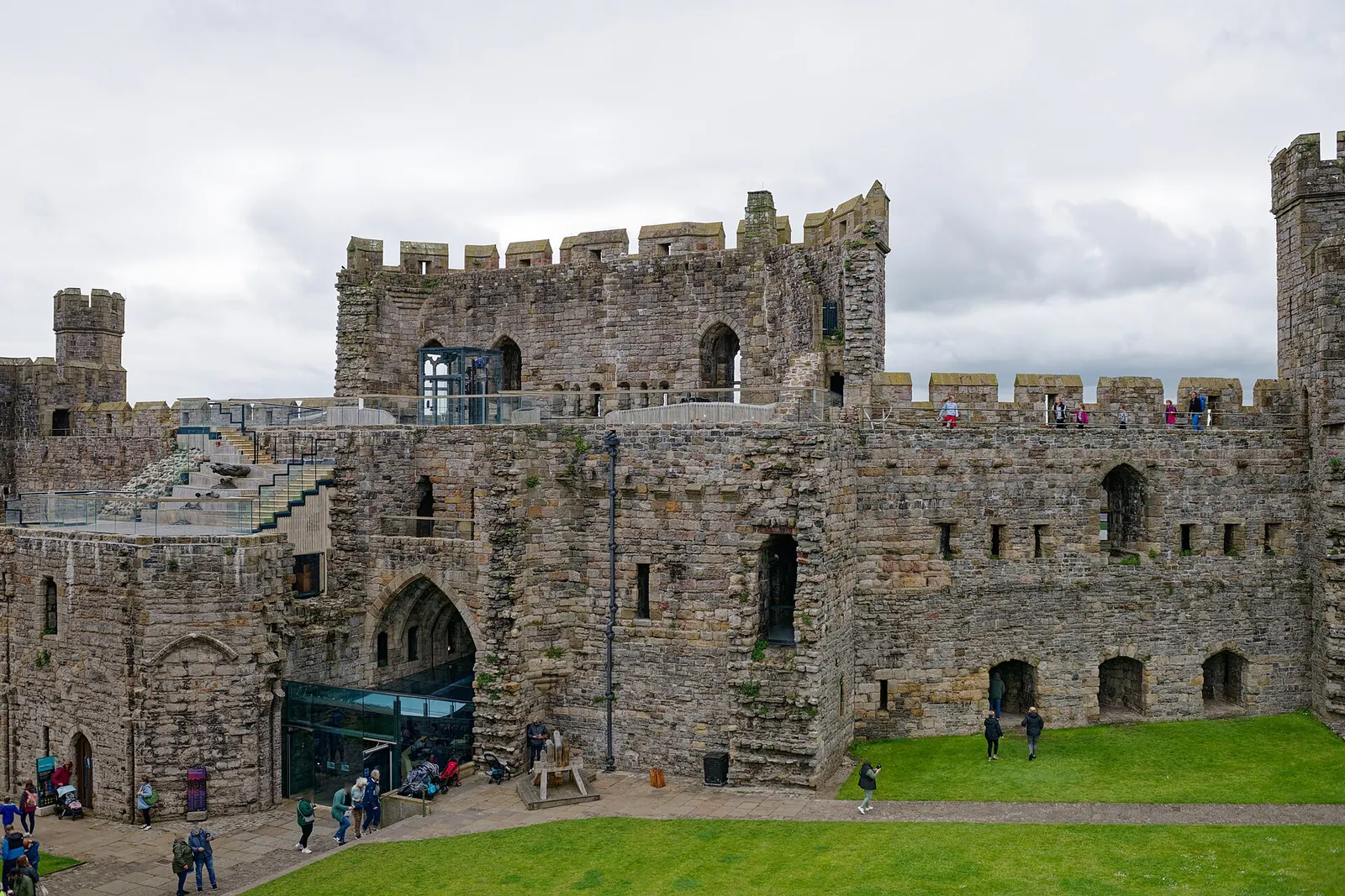 King's Gate at Caernarfon Castle, seen from within the walls: a modern glass lift and stone stairway lead up to the crenellated wall-walk, visitors small along the battlements.