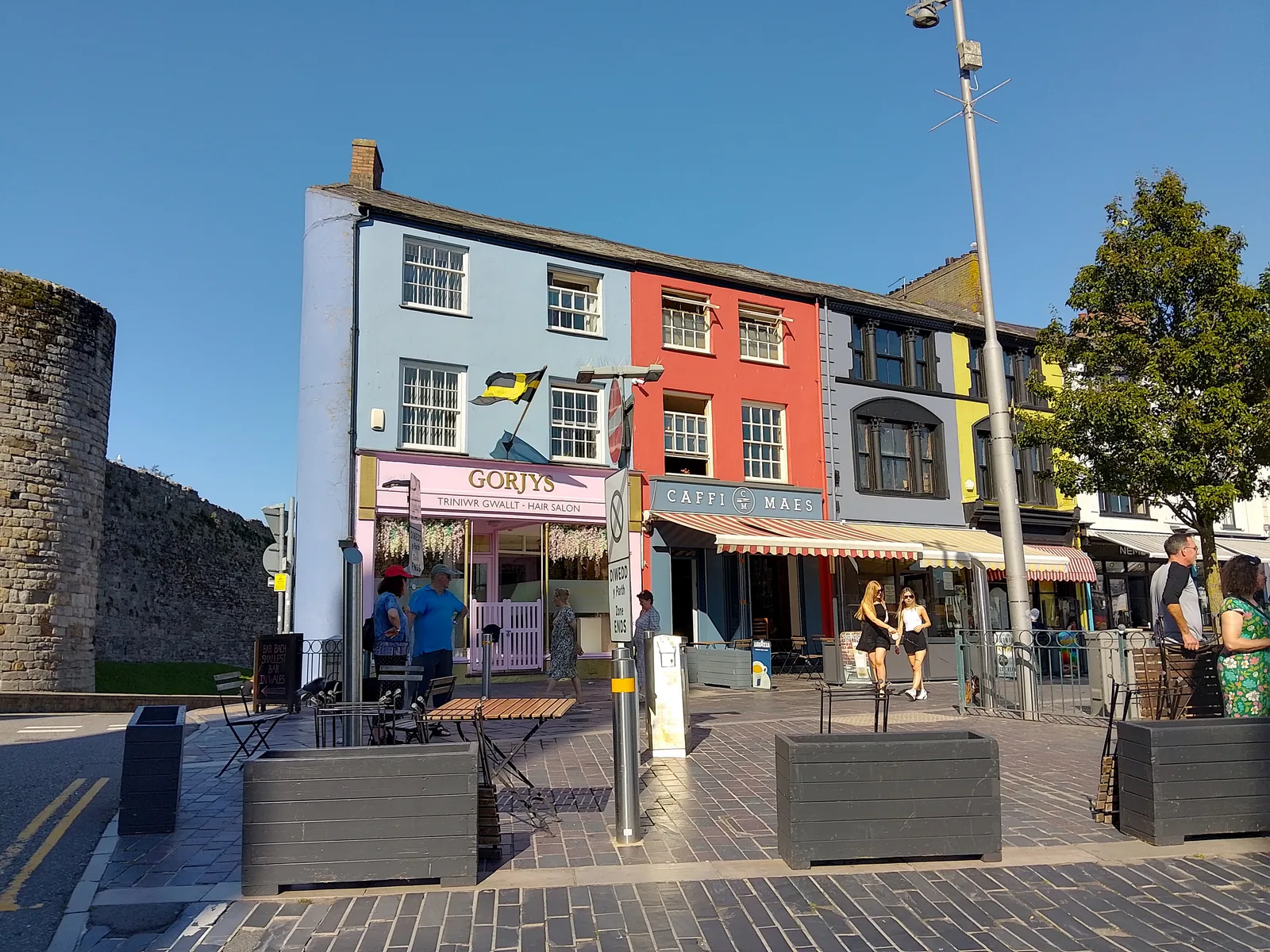Painted shopfronts and pavement tables on the east side of Y Maes, Caernarfon, with the town wall at the left edge