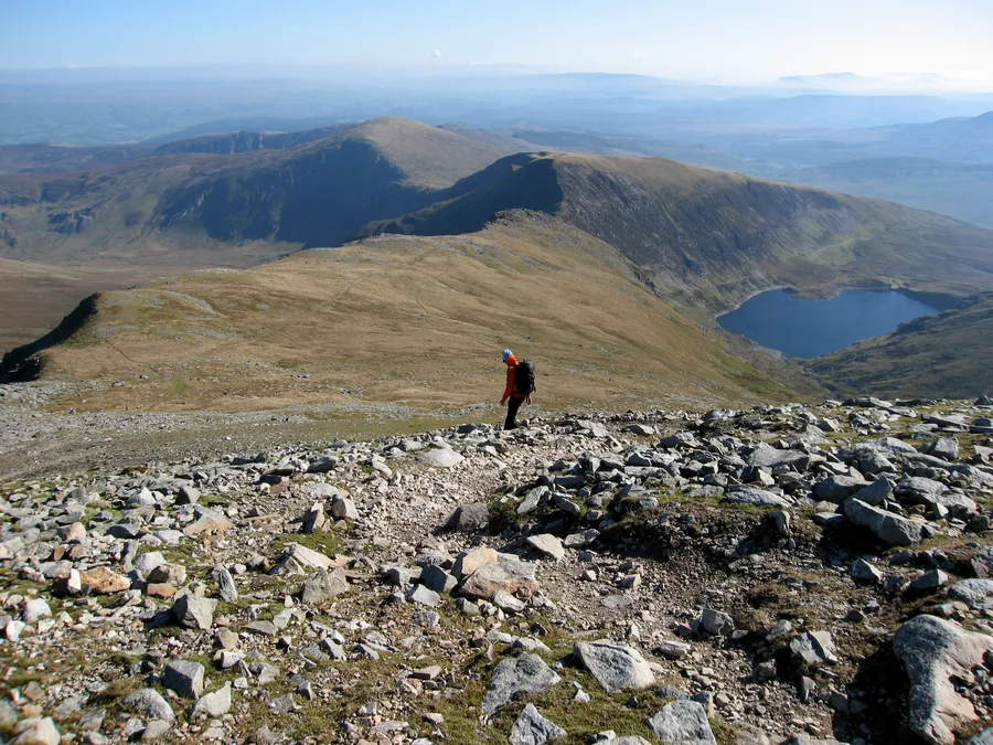 A walker on the stony ridge below the summit of Carnedd Llewelyn, a mountain tarn in the cwm and the Carneddau running on beyond.