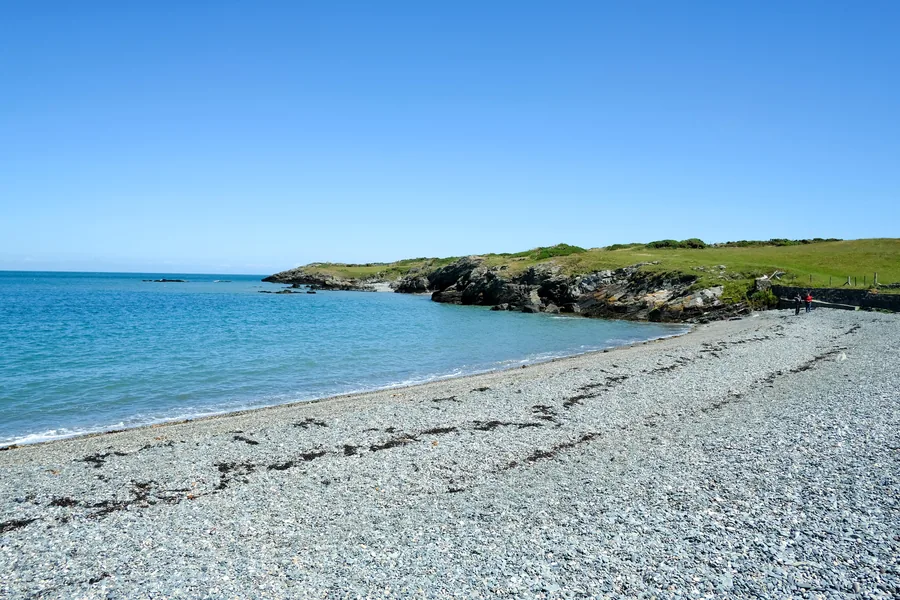 The shingle ridge of Esgair Cemlyn curving round Cemlyn Bay under a clear sky, turquoise water on the seaward side.