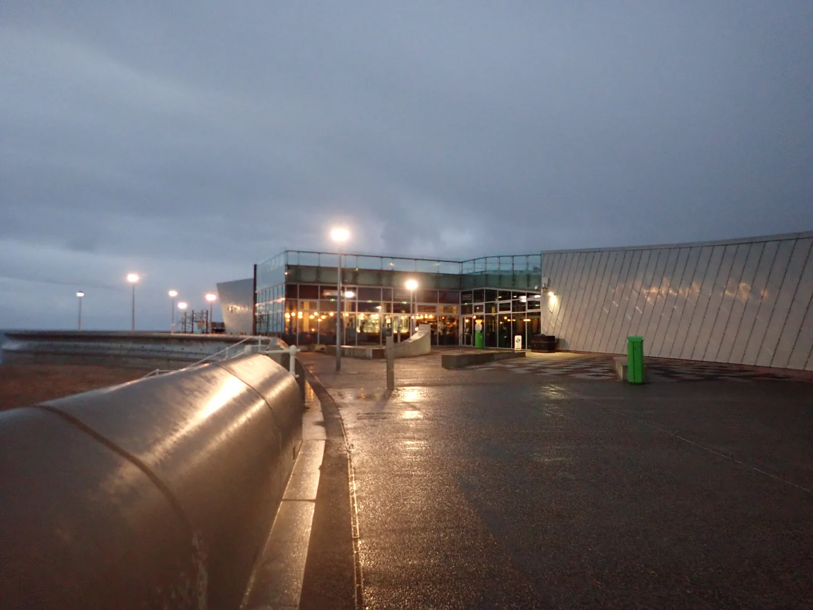 The Porth Eirias watersports building lit at dusk, its glass frontage reflected in the wet promenade