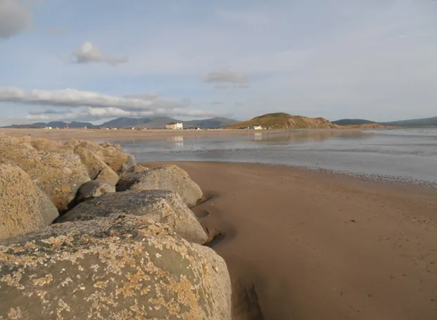 Rocks in the foreground of Dinas Dinlle beach, with the tide out and hills across the water in the distance.