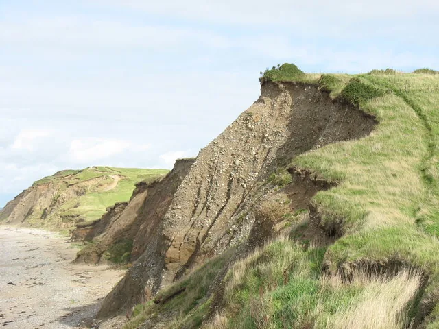 A dramatically eroding clay cliff face at Dinas Dinlle, layered soil and stone crumbling toward the beach below.