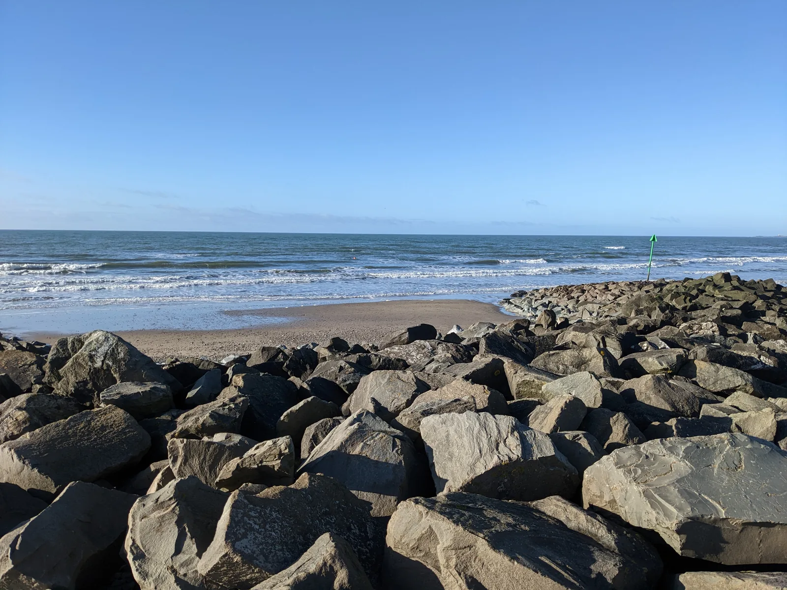 Large weathered rocks of a rocky spur at Dinas Dinlle, splitting the beach in two, with the sea and a channel marker beyond.