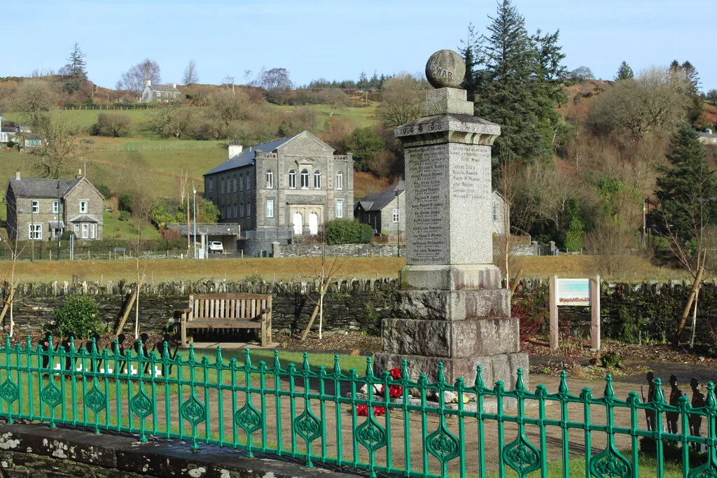 The war memorial at Dolwyddelan, a stone column behind green railings with the village houses rising behind
