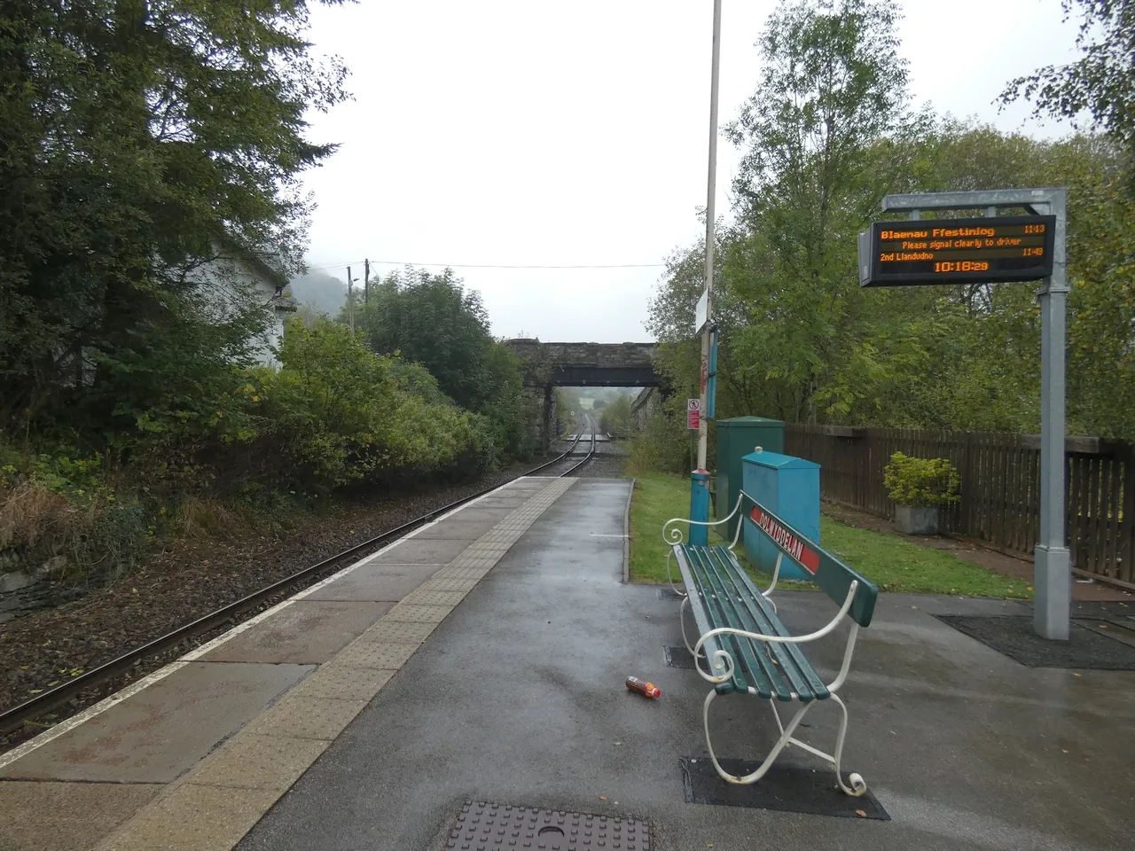 The single platform at Dolwyddelan station, a bench and an information display beside the line under a road bridge