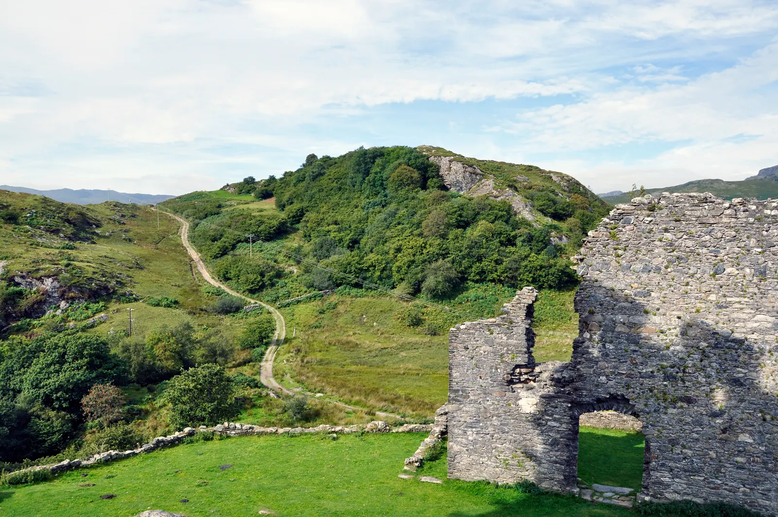 The Lledr valley seen from Dolwyddelan Castle, a path running down past ruined walling towards green fields and forested slopes