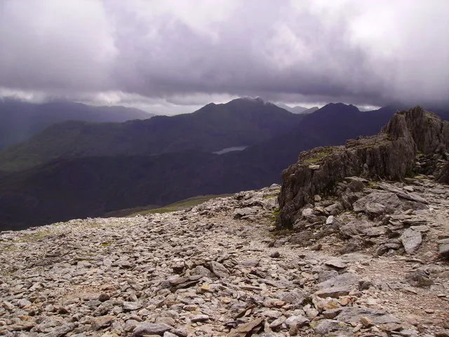 The shattered rock plateau of Glyder Fawr under heavy cloud, with a weathered tor on the right and a long mountain ridge on the skyline across the valley