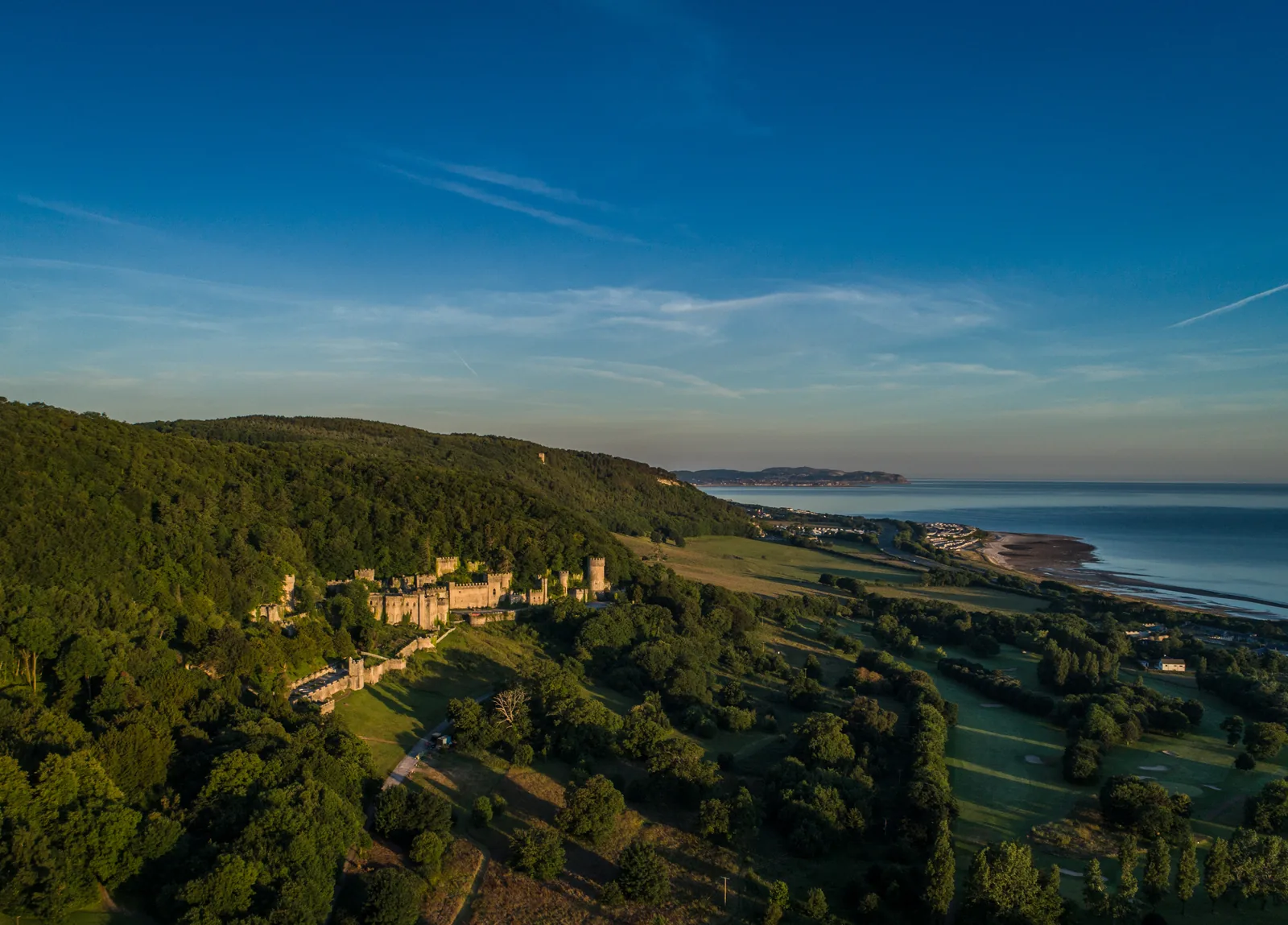 A golden-hour aerial view of the Gwrych Castle estate above the North Wales coast, the sea visible in the distance.