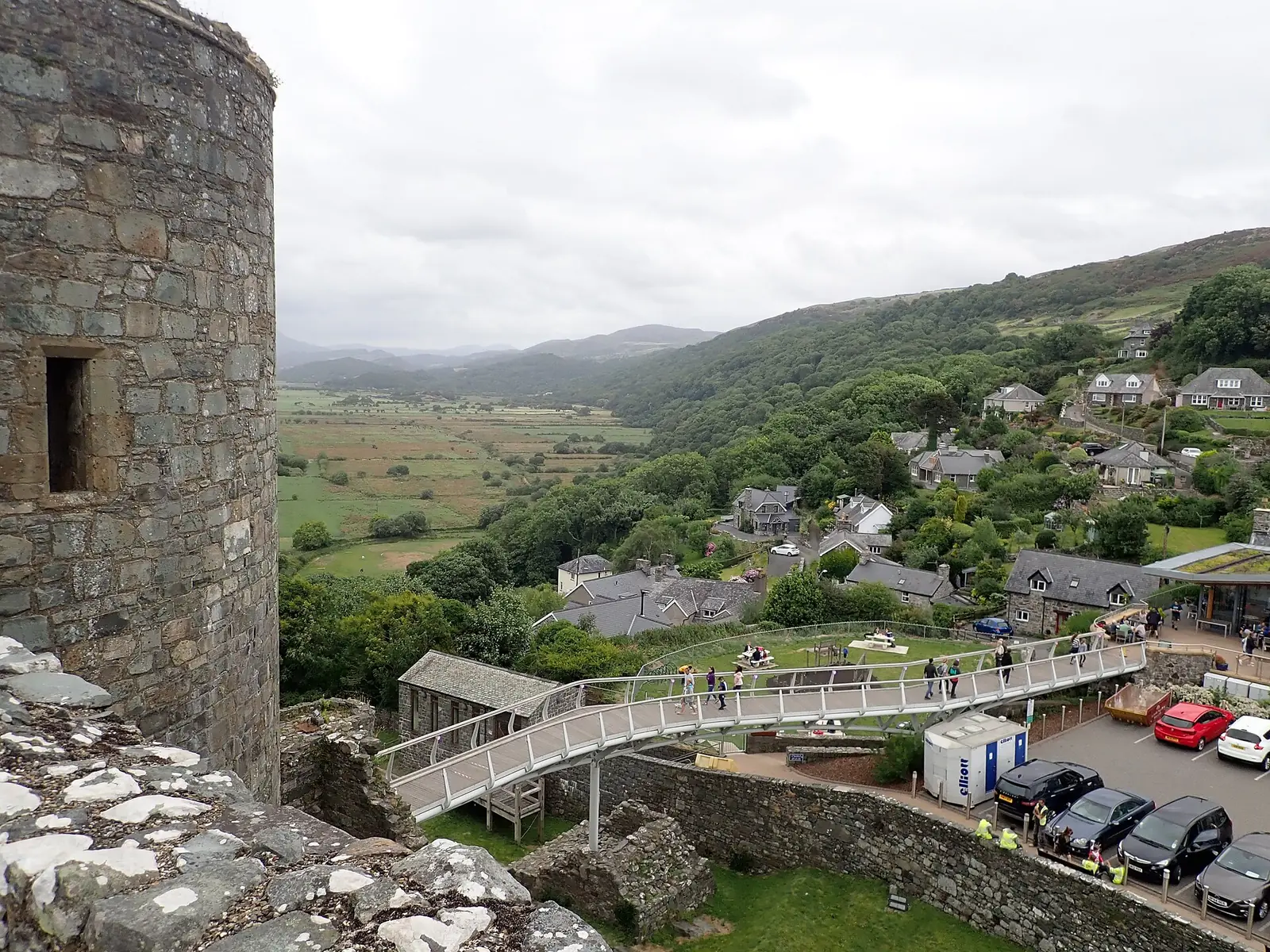 The modern footbridge into Harlech Castle, a white walkway leading down to the visitor centre and car park, a few visitors crossing, green hills behind.