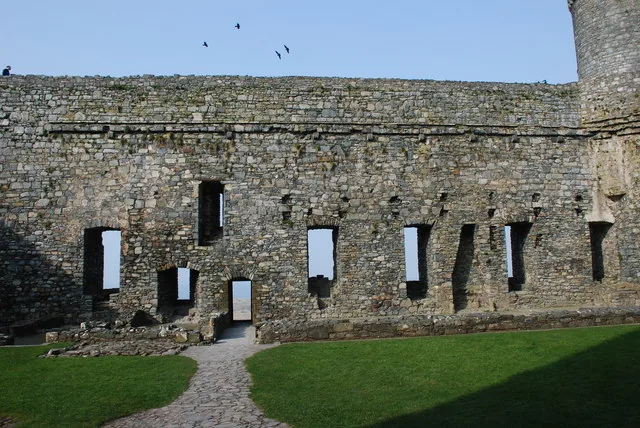 A stone curtain wall inside Harlech Castle's ward, arched window openings along its length, a cobbled path and green lawn below, birds overhead.