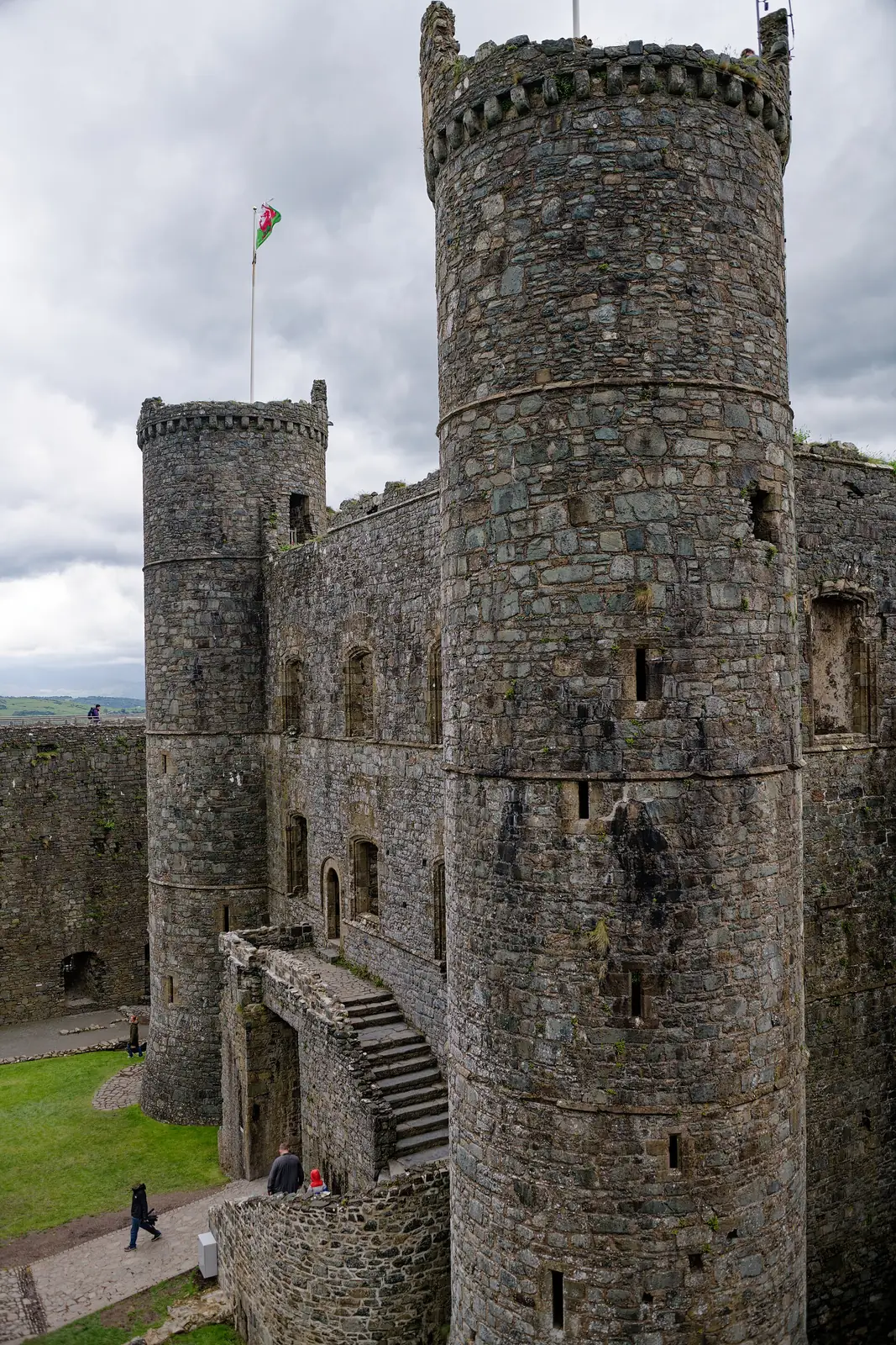 Two large round towers of Harlech Castle's gatehouse, seen close from the wall-walk, a Welsh flag flying above and small figures on the battlements below.