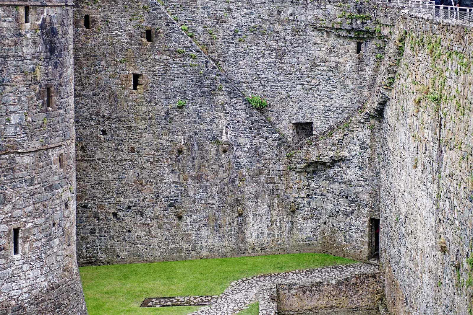 Weathered stone walls inside Harlech Castle's south-east corner, seen from the wall-walk, a strip of green lawn below.