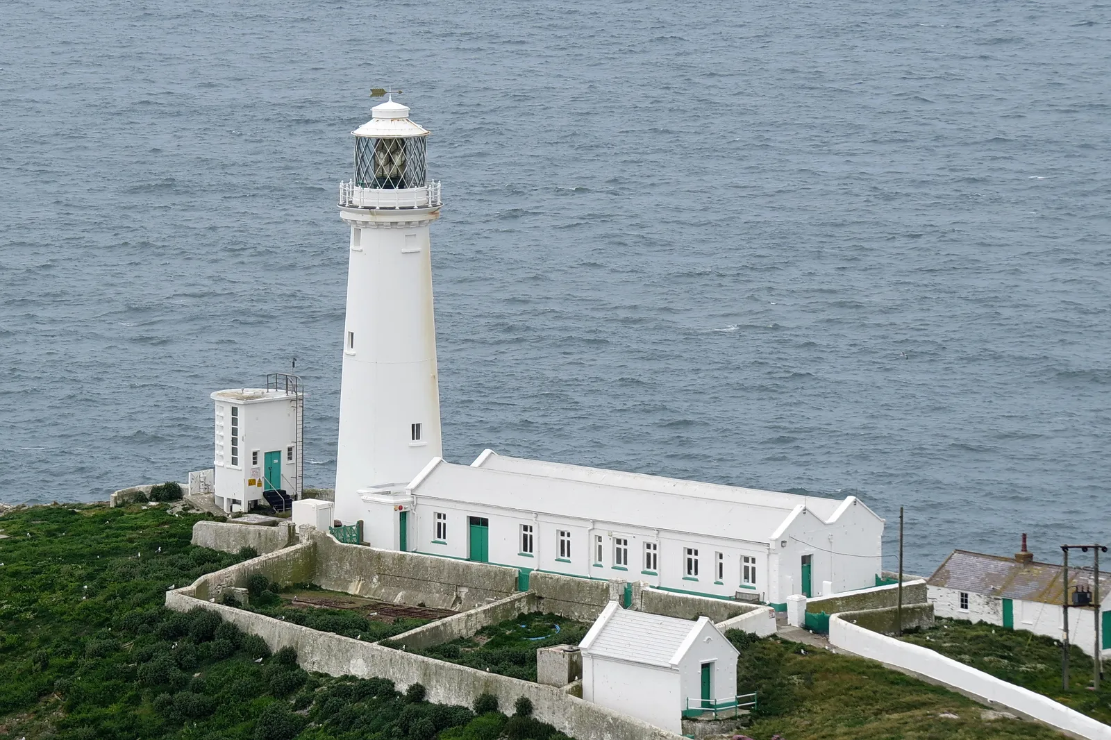 South Stack Lighthouse on its island below the cliffs of Holy Island, seen from the headland above — the lighthouse and its bridge in clear coastal light.