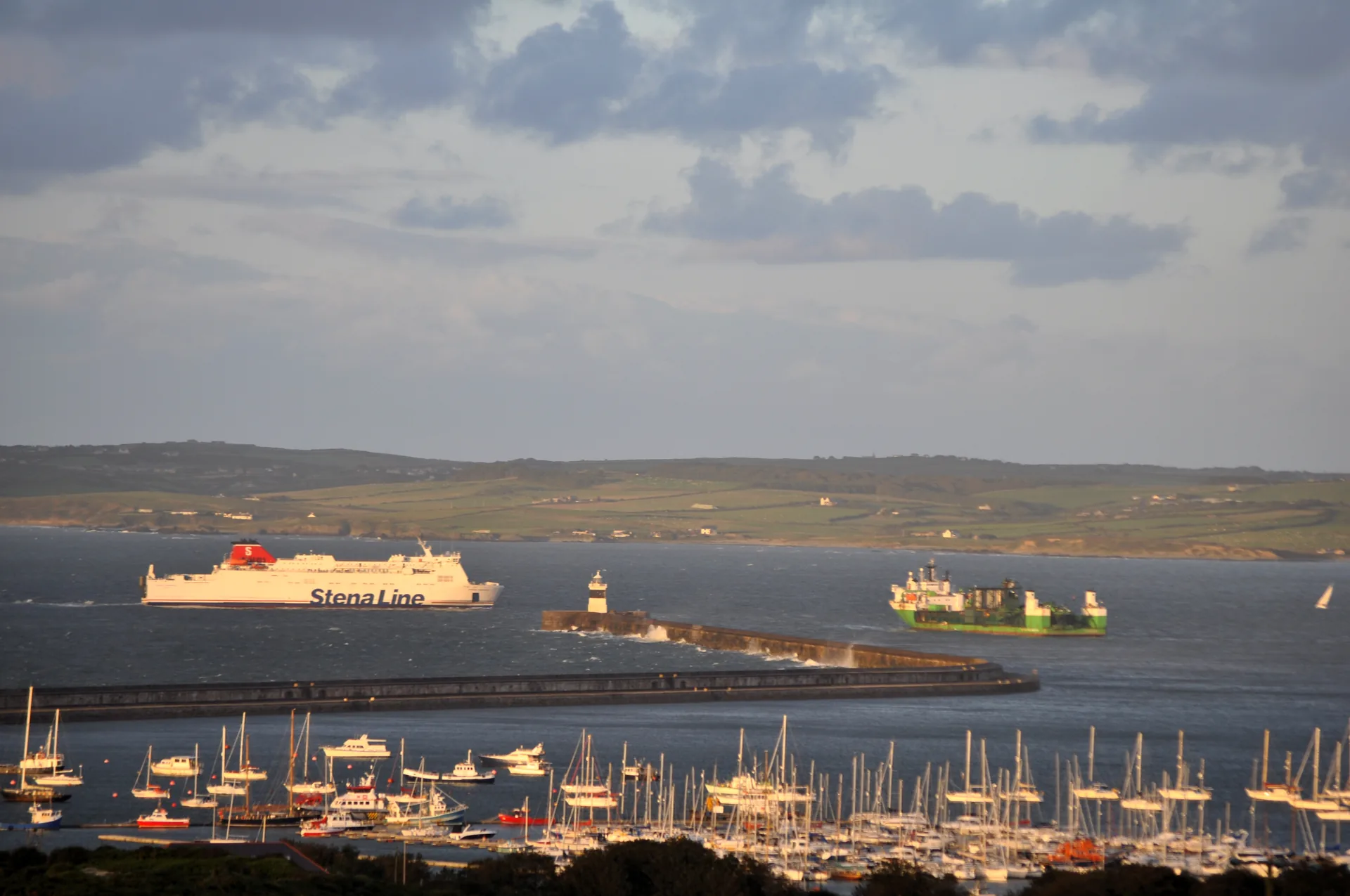 Holyhead harbour in low evening sun: a Stena Line ferry standing off the breakwater and its lighthouse, a cargo ship beyond, and the marina full of masts in the foreground