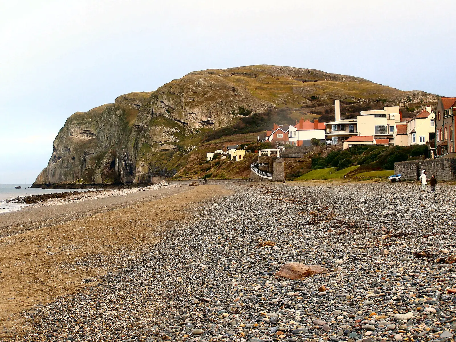 The Little Orme headland seen from the shingle beach at Ormes Bay, with houses along the shore below the cliff.