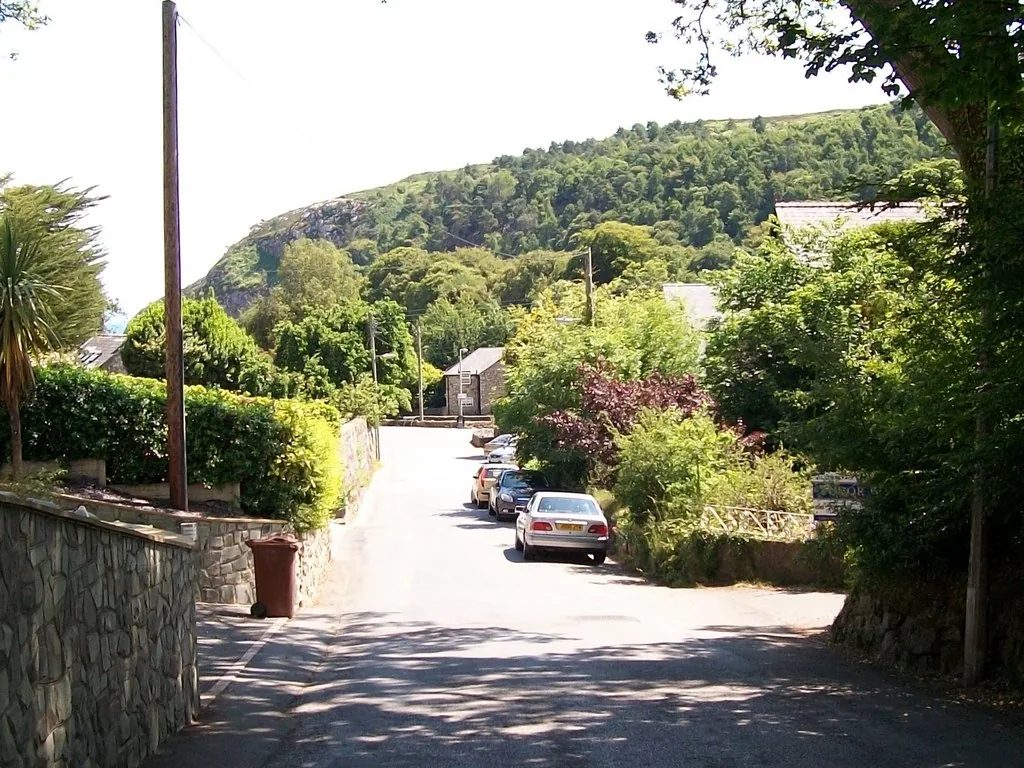 The lane through Llanbedrog running down between stone walls and hedges, the wooded headland rising behind