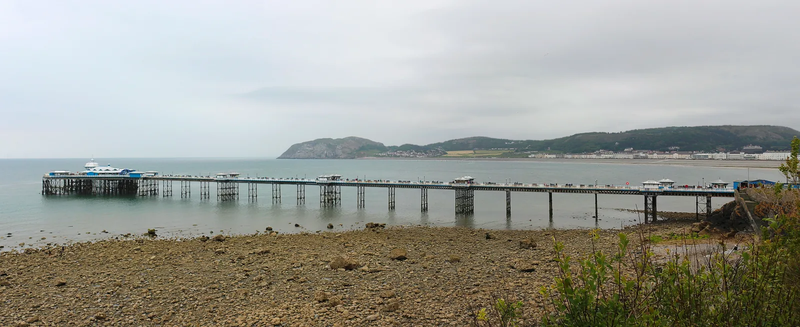 The full length of Llandudno Pier at low tide, its ironwork legs standing in shallow water with the Little Orme beyond