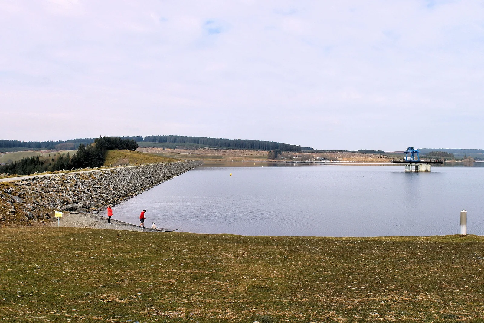 The rock-armoured face of Brenig Dam with the draw-off tower standing out in the lake, and two walkers on the shore for scale.