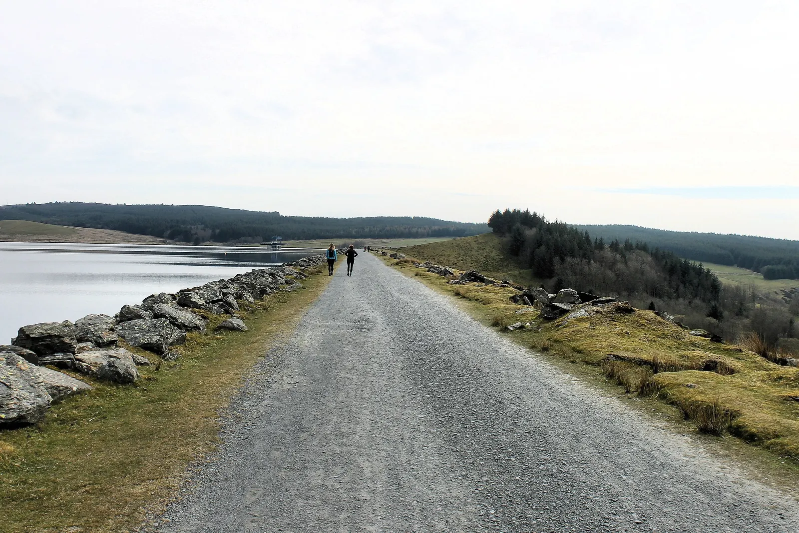 Walkers on the surfaced gravel track along the crest of Brenig Dam, with rock armour on one side and moorland on the other.
