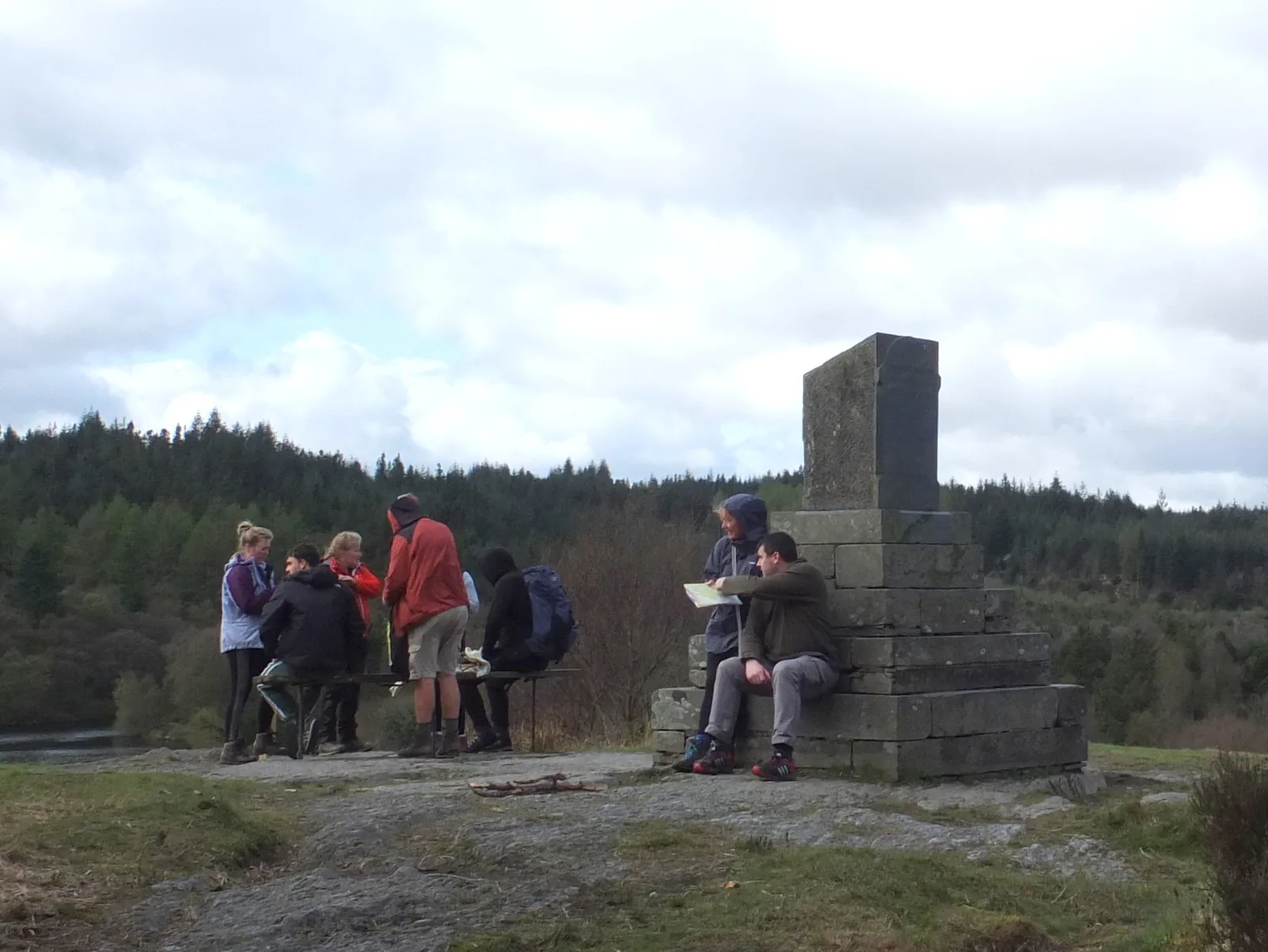 The stone memorial above Llyn Elsi, a tall block on a stepped plinth, with walkers resting at its base and forest behind.